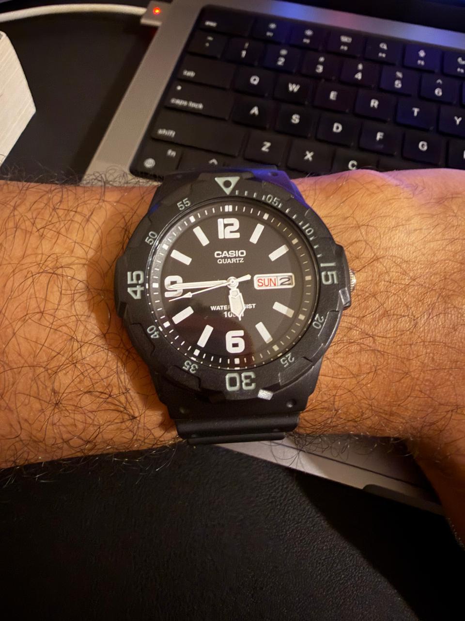 A black Casio quartz watch with a black dial worn on a wrist, resting against a laptop keyboard.