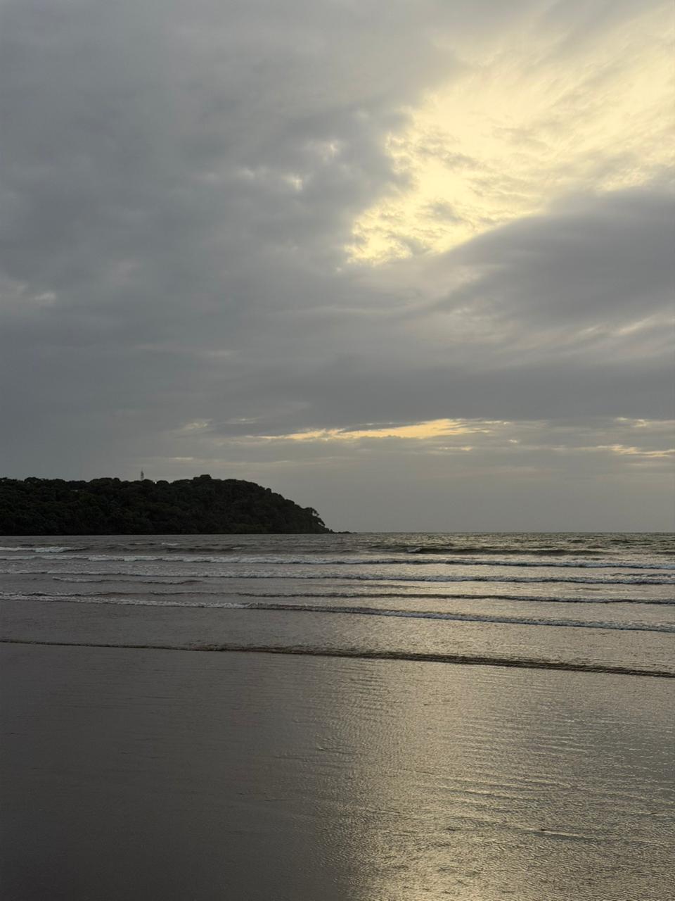 Photo of Anjuna Beach at dusk, with grey clouds over the sea, gentle waves rolling in, and a tree-covered headland silhouetted on the left.