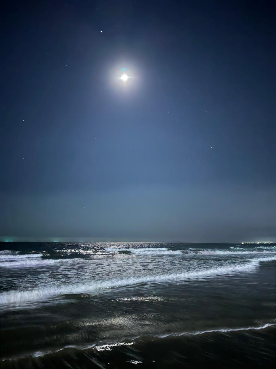 Photo of Anjuna Beach at night, with a bright moon in a dark blue sky and white-capped waves rolling toward the shore.